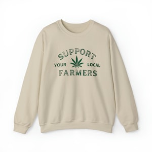 Puede incluir: Una sudadera beige con el texto "SUPPORT YOUR LOCAL FARMERS" impreso en la parte delantera. Una hoja de marihuana verde est&aacute; en el centro del texto.