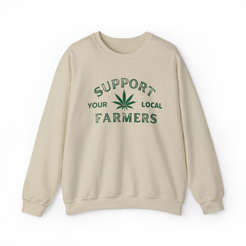 Puede incluir: Una sudadera beige con el texto "SUPPORT YOUR LOCAL FARMERS" impreso en la parte delantera. Una hoja de marihuana verde est&aacute; en el centro del texto.