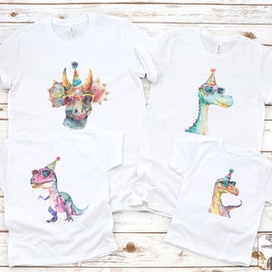 May include: Four white t-shirts with watercolor dinosaur designs. Each dinosaur wears sunglasses and a party hat. The dinosaurs are a triceratops, a brontosaurus, a T-Rex, and a raptor. The shirts are on a light wood background.