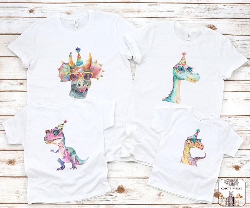 May include: Four white t-shirts with watercolor dinosaur designs. Each dinosaur wears sunglasses and a party hat. The dinosaurs are a triceratops, a brontosaurus, a T-Rex, and a raptor. The shirts are on a light wood background.