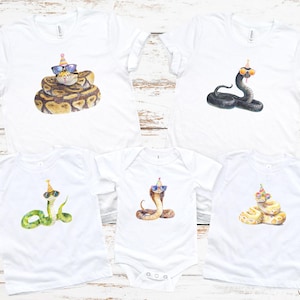 May include: White t-shirts and a onesie featuring watercolor illustrations of snakes wearing sunglasses and party hats. The snakes vary in color and pattern, with designs including a ball python, cobra, and other snake species.
