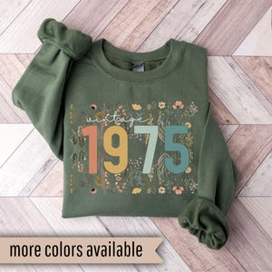 Puede incluir: Una sudadera verde con un dise&ntilde;o floral y el texto "vintage 1975" en letras de colores.