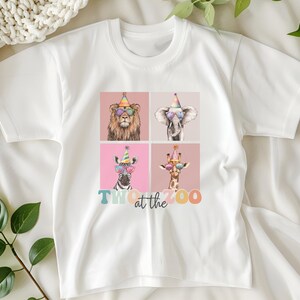 Op de afbeelding: Wit T-shirt met een afbeelding van vier dieren die feestmutsen en zonnebrillen dragen. De tekst "Two at the Zoo" is onder de dieren gedrukt.