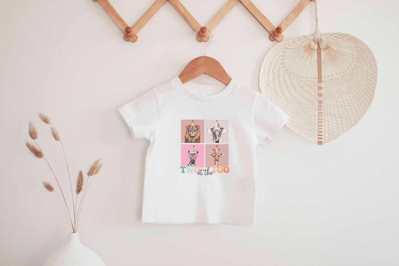 Op de afbeelding: Wit T-shirt met een kleurrijke afbeelding van dieren die feestmutsen dragen. De tekst "Party at the Zoo" is onder de afbeelding gedrukt.