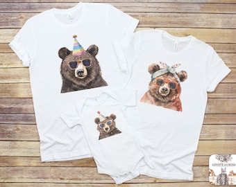 May include: A set of three white t-shirts featuring watercolor illustrations of bears wearing sunglasses. The smallest shirt has a bear wearing a party hat, the medium shirt has a bear wearing a bandana, and the largest shirt has a bear wearing sunglasses.