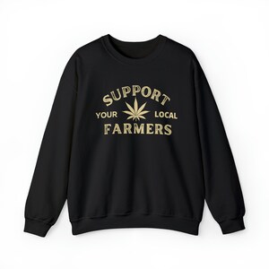Puede incluir: Sudadera negra con un gr&aacute;fico dorado que dice "Apoya a tus agricultores locales" con una hoja de marihuana en el centro.