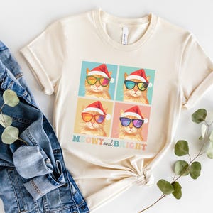 May include: Cream-colored t-shirt featuring four squares with illustrations of orange cats wearing Santa hats and sunglasses. The text "MEOWY and BRIGHT" is below the squares. The shirt is paired with a denim jacket and a sprig of greenery.