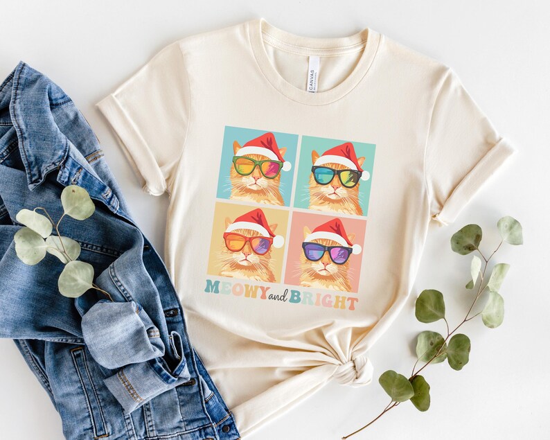 May include: Cream-colored t-shirt featuring four squares with illustrations of orange cats wearing Santa hats and sunglasses. The text "MEOWY and BRIGHT" is below the squares. The shirt is paired with a denim jacket and a sprig of greenery.