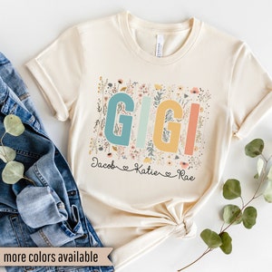 May include: A beige t-shirt with a floral design and the word "GIGI" in colorful letters. The names "Jacob", "Katie", and "Rae" are written below the word "GIGI".