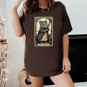 May include: A brown t-shirt with a graphic of a tarot card featuring a mouse dressed as a hermit. The card is titled "The Hermit".