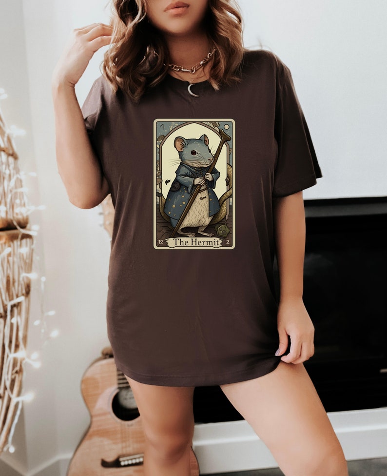 May include: A brown t-shirt with a graphic of a tarot card featuring a mouse dressed as a hermit. The card is titled "The Hermit".