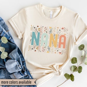 Op de afbeelding: Een cr&egrave;mekleurig T-shirt met het woord "NANA" in regenboogkleuren omringd door een bloemenontwerp.