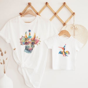 May include: Two white t-shirts, one adult and one child size, each featuring a colorful watercolor dinosaur design. The larger shirt has a triceratops wearing sunglasses and a party hat. The smaller shirt has a pterodactyl with a party hat.