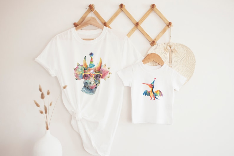 May include: Two white t-shirts, one adult and one child size, each featuring a colorful watercolor dinosaur design. The larger shirt has a triceratops wearing sunglasses and a party hat. The smaller shirt has a pterodactyl with a party hat.