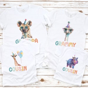 May include: Four white t-shirts with colorful animal graphics and text. The shirts feature a hyena, an ostrich, a giraffe, and a hippopotamus. The text on the shirts reads "Grandpa", "Grammy", "Cousin", and "Cousin".