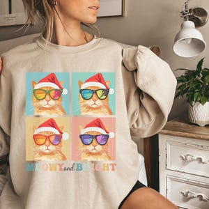 May include: Beige sweatshirt featuring a colorful graphic of four cats wearing Santa hats and sunglasses. The cats have different colored sunglasses. The text "MEOWY and BRIGHT" is below the graphic.