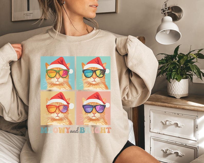 May include: Beige sweatshirt featuring a colorful graphic of four cats wearing Santa hats and sunglasses. The cats have different colored sunglasses. The text "MEOWY and BRIGHT" is below the graphic.
