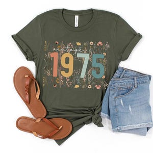 Puede incluir: Una camiseta verde con un dise&ntilde;o floral y el texto "vintage 1975" en letras de colores.