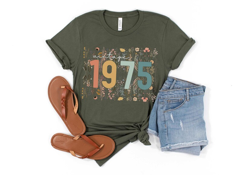 Puede incluir: Una camiseta verde con un dise&ntilde;o floral y el texto "vintage 1975" en letras de colores.
