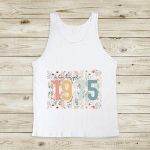 Puede incluir: Camiseta sin mangas blanca con un dise&ntilde;o floral y el texto "vintage 1975" en letras de colores.
