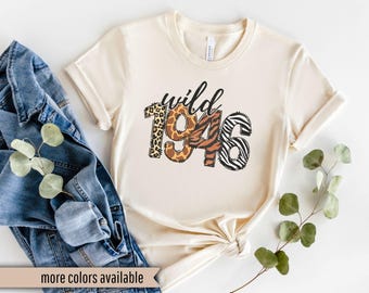 May include: Cream-colored t-shirt with the word "wild" in black script and the numbers "1946" in animal print patterns. The shirt is styled with rolled-up sleeves and a knot at the bottom. A denim jacket and greenery are in the background.