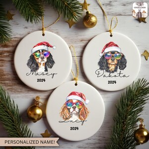 May include: Three white ceramic Christmas ornaments featuring Cavalier King Charles Spaniels wearing Santa hats and sunglasses. Each ornament has a different name and the year 2024 printed below the dog's portrait. Gold string for hanging.
