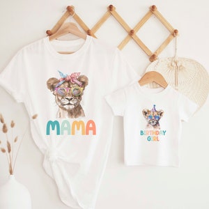 May include: Two white t-shirts with a lion graphic. The larger shirt says "MAMA" in colorful letters. The smaller shirt says "BIRTHDAY GIRL" in colorful letters.