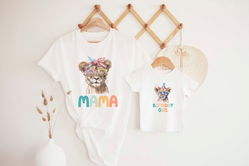 May include: Two white t-shirts with a lion graphic. The larger shirt says "MAMA" in colorful letters. The smaller shirt says "BIRTHDAY GIRL" in colorful letters.