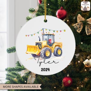 May include: White ceramic ornament featuring a yellow tractor with a front loader, decorated with colorful lights. The name "Tyler" and the year "2024" are written in black script. The ornament hangs on a Christmas tree.