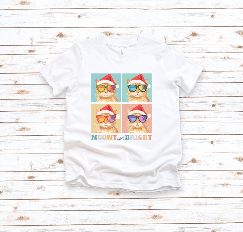 May include: White t-shirt featuring a colorful graphic of four cats wearing Santa hats and sunglasses. The cats have different colored lenses. The text "MEOWY and BRIGHT" is printed below the graphic. The shirt is displayed on a light wood background.