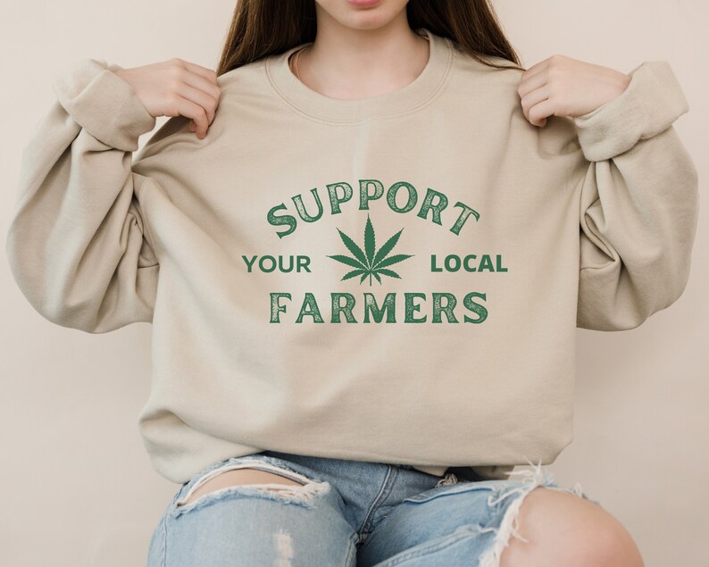 Puede incluir: Una sudadera beige con un gr&aacute;fico de hoja de marihuana verde y el texto "Apoya a tus agricultores locales".