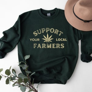 Puede incluir: Una sudadera verde oscuro con el texto "Support Your Local Farmers" en fuente dorada. Un gr&aacute;fico de hoja de marihuana est&aacute; en el centro del texto.
