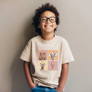Pu&ograve; includere: Un giovane indossa una maglietta beige con una stampa grafica di quattro animali che indossano cappelli da festa e occhiali da sole. Gli animali sono un leone, un elefante, una zebra e una giraffa.