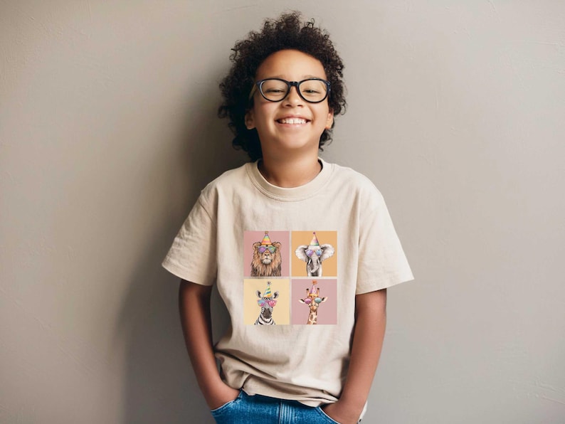 Pu&ograve; includere: Un giovane indossa una maglietta beige con una stampa grafica di quattro animali che indossano cappelli da festa e occhiali da sole. Gli animali sono un leone, un elefante, una zebra e una giraffa.