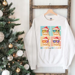 May include: White sweatshirt featuring a colorful graphic of four cats wearing Santa hats and sunglasses. The text "MEOWY and BRIGHT" is printed below the cats in a rainbow font. The sweatshirt is hanging on a wooden rack near a Christmas tree.