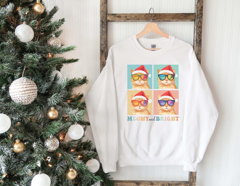 May include: White sweatshirt featuring a colorful graphic of four cats wearing Santa hats and sunglasses. The text "MEOWY and BRIGHT" is printed below the cats in a rainbow font. The sweatshirt is hanging on a wooden rack near a Christmas tree.