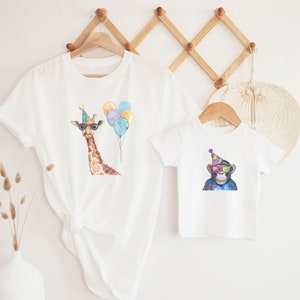 May include: Two white t-shirts with animal illustrations. The larger shirt features a giraffe wearing a party hat and sunglasses, with colourful balloons. The smaller shirt features a monkey wearing a party hat and sunglasses.