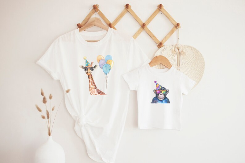 May include: Two white t-shirts with animal illustrations. The larger shirt features a giraffe wearing a party hat and sunglasses, with colourful balloons. The smaller shirt features a monkey wearing a party hat and sunglasses.