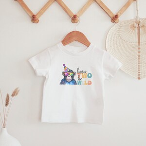 May include: White baby t-shirt featuring a colourful illustration of a monkey wearing sunglasses and a party hat. The text "born two be wild" is printed on the shirt.