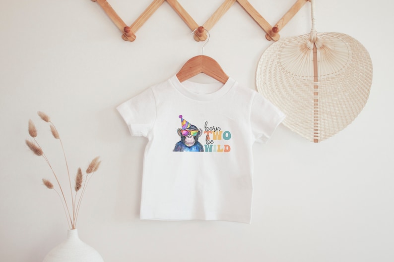 May include: White baby t-shirt featuring a colourful illustration of a monkey wearing sunglasses and a party hat. The text "born two be wild" is printed on the shirt.