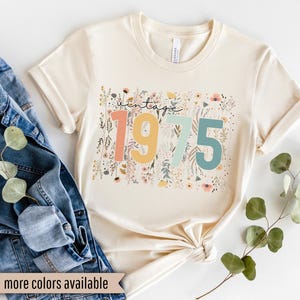 Puede incluir: Una camiseta beige con un dise&ntilde;o floral y el texto "vintage 1975" en letras de colores.
