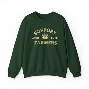 Puede incluir: Una sudadera verde oscuro con el texto "Support Your Local Farmers" en letras doradas. Una hoja de marihuana estilizada est&aacute; en el centro del texto.