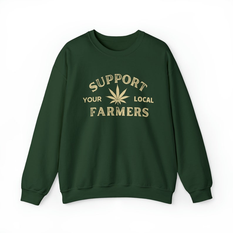 Puede incluir: Una sudadera verde oscuro con el texto "Support Your Local Farmers" en letras doradas. Una hoja de marihuana estilizada est&aacute; en el centro del texto.