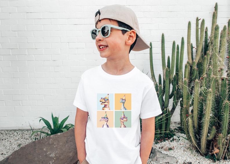 Puede incluir: Una camiseta blanca con un gr&aacute;fico de cuatro dinosaurios que llevan sombreros de fiesta y gafas de sol. Los dinosaurios est&aacute;n en un patr&oacute;n de cuadr&iacute;cula en la camiseta. La camiseta la lleva un ni&ntilde;o que lleva una gorra de b&eacute;isbol beige y gafas de sol azules.