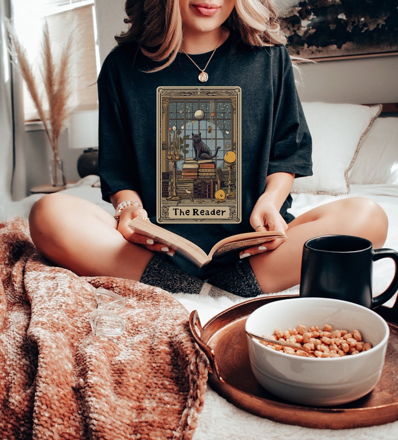 May include: A black t-shirt featuring a vintage tarot card design with a black cat sitting on a stack of books. The card is titled "The Reader".