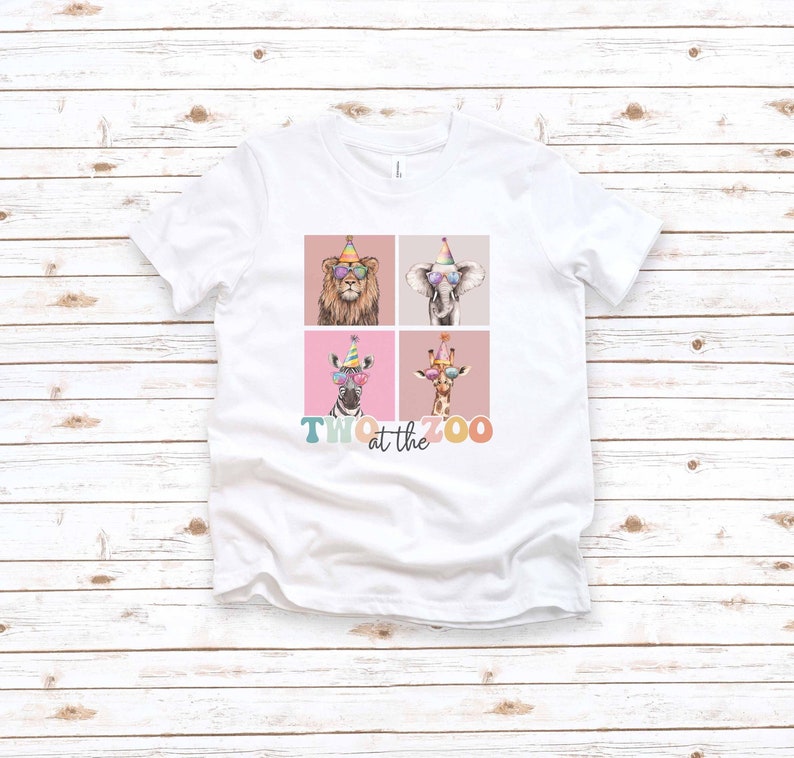 Op de afbeelding: Wit T-shirt met een afbeelding van vier dieren die feestmutsen en zonnebrillen dragen. De dieren zijn een leeuw, een olifant, een zebra en een giraffe. De tekst "TWO at the ZOO" staat onder de dieren.
