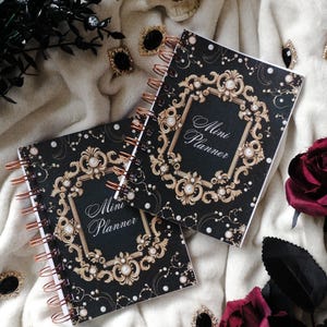 Puede incluir: Dos Mini Planners negros con detalles dorados y perlas y encuadernación en espiral. La portada presenta un marco decorativo con las palabras "Mini Planner" en escritura elegante. Los planificadores están sobre una superficie blanca y suave con elementos decorativos.