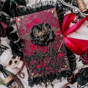 Op de afbeelding: Een rood fluwelen dagboek met zwarte kantversiering en een gouden metalen vleermuisbedel. Het dagboek is versierd met een zwarte vleermuis en een gouden sleutel.