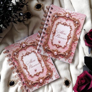 May include: Two spiral-bound "Mini Planner" notebooks with pink covers featuring ornate gold frames, pearls, and lace details. The notebooks are surrounded by decorative elements, including black jewelry, a skull, and dark red roses.