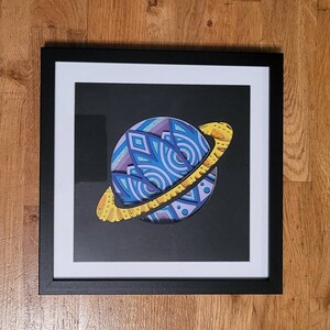 May include: Framed art print featuring a layered paper design of a planet with a ring. The planet is primarily blue and purple with geometric patterns. The ring is yellow with a textured design. The artwork is set against a black background and framed in a black frame.